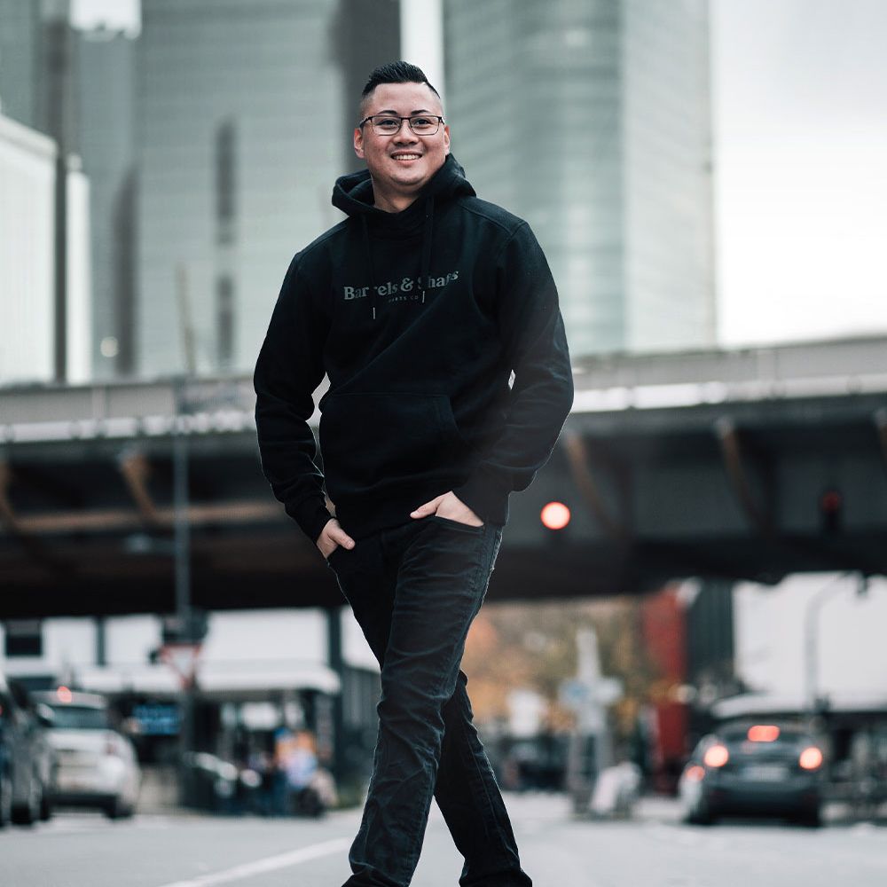 Barrels and Shafts Hoodie - Schwarz Eine Person trägt einen schwarzen "Barrels and Shafts" Hoodie und steht entspannt auf einer Straße. Im Hintergrund sind unscharfe Gebäude und Autos zu sehen.