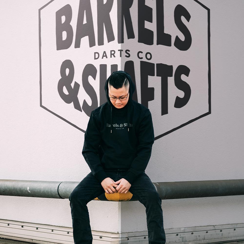 Barrels and Shafts Hoodie - Schwarz Eine Person trägt einen schwarzen "Barrels and Shafts" Hoodie und sitzt auf einer Bank. Im Hintergrund ist ein großes Wandlogo mit der Aufschrift "Barrels & Shafts Darts Co" zu sehen.
