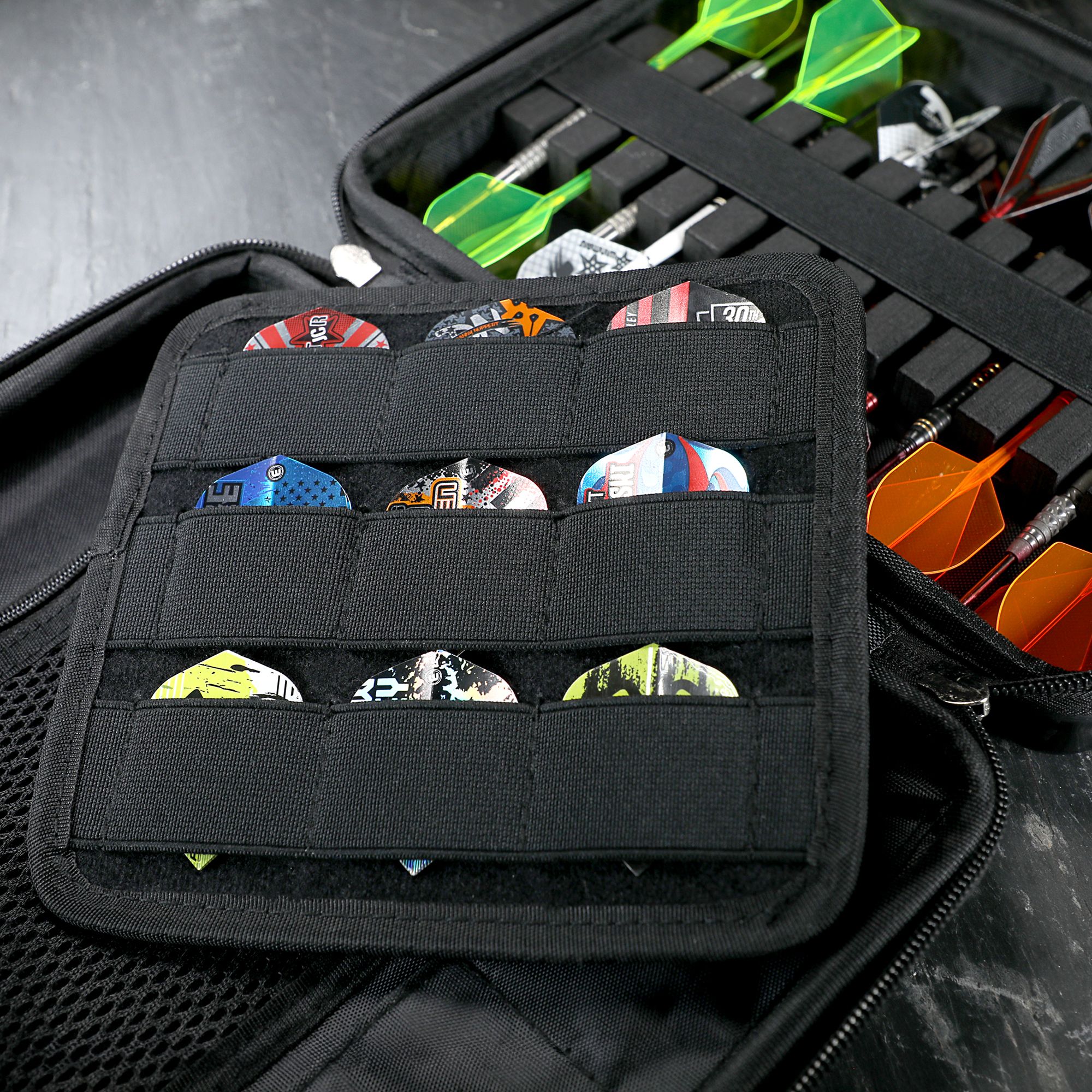 Das Bild zeigt ein geöffnetes Dart-Etui mit Fächern für verschiedene Dart-Flights. Im Hintergrund sind mehrere vollständig montierte Darts mit bunten Flights zu sehen.
