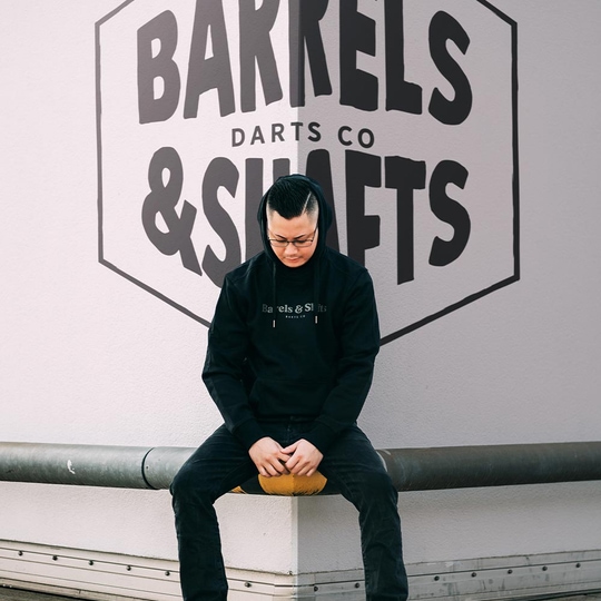 Barrels and Shafts Hoodie - Schwarz Eine Person trägt einen schwarzen "Barrels and Shafts" Hoodie und sitzt auf einer Bank. Im Hintergrund ist ein großes Wandlogo mit der Aufschrift "Barrels & Shafts Darts Co" zu sehen.